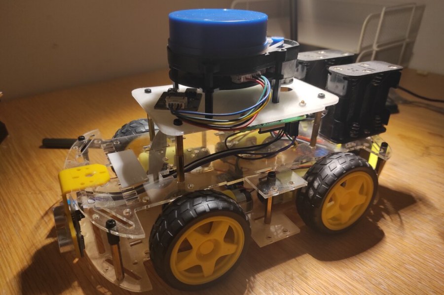 The four-wheeled rover on a desk: a clear acrylic chassis, yellow-hubbed wheels, a blue lidar puck on the upper deck and two battery packs at the back.